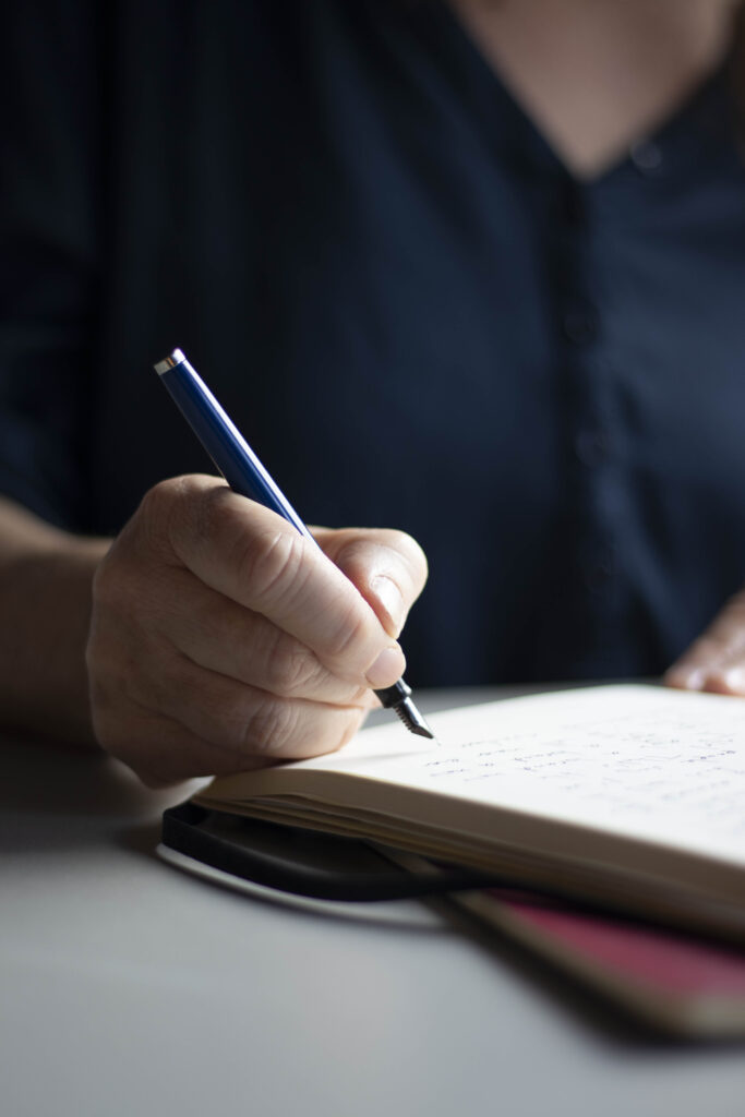 Een hand (de mijne) met een blauwe vulpen, schrijvend in een notitieboek.